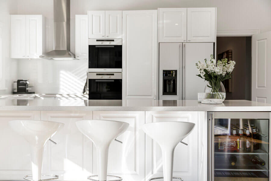 High-gloss white kitchen with a peninsula, white moulded bar stools, integrated wine fridge and built-in double ovens